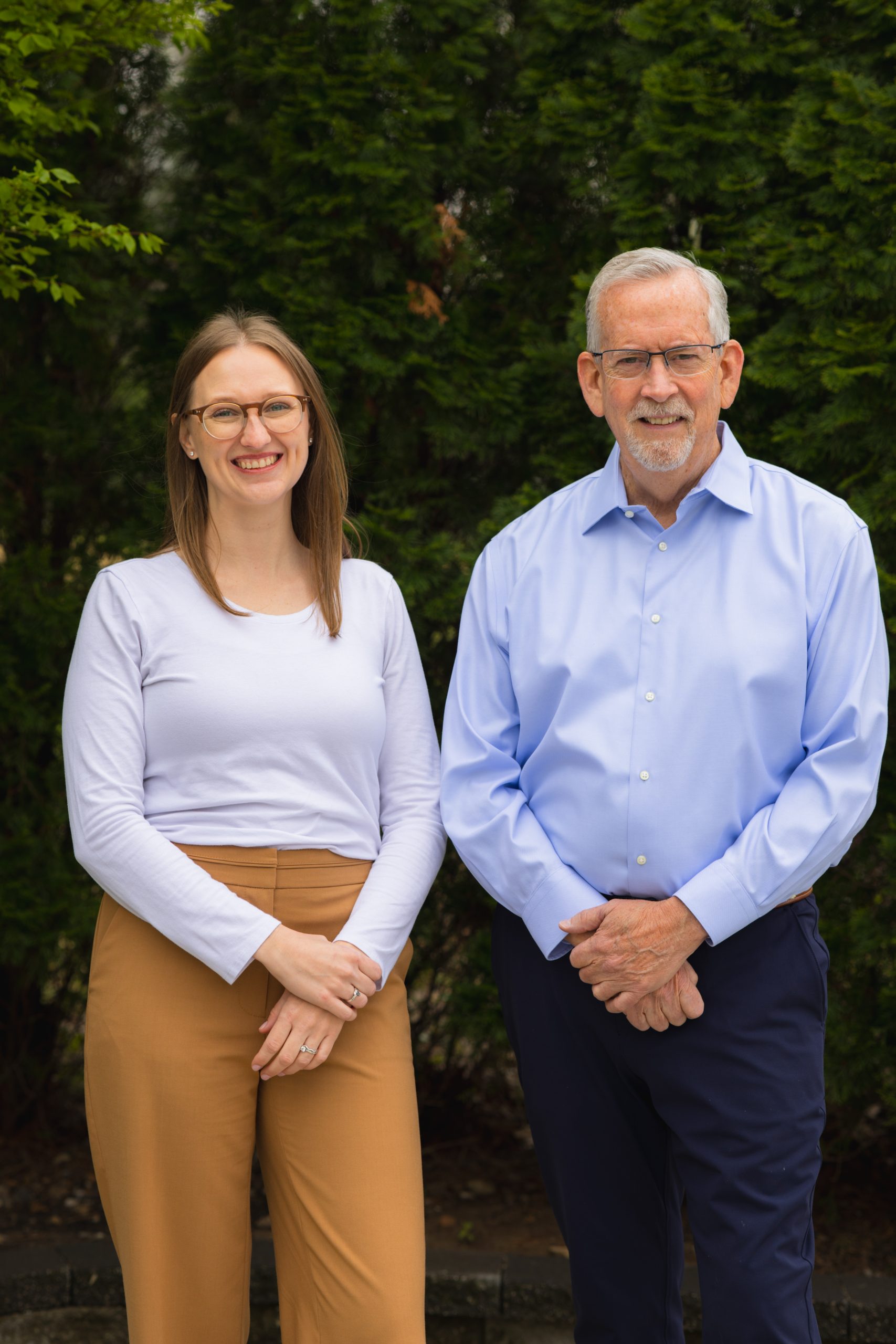 A photo of Alyssa Clarkin and Dan Clarkin standing in front of some shrubs. They are both standing with their arms in front of them, Alyssa on the left and Dan on the right.